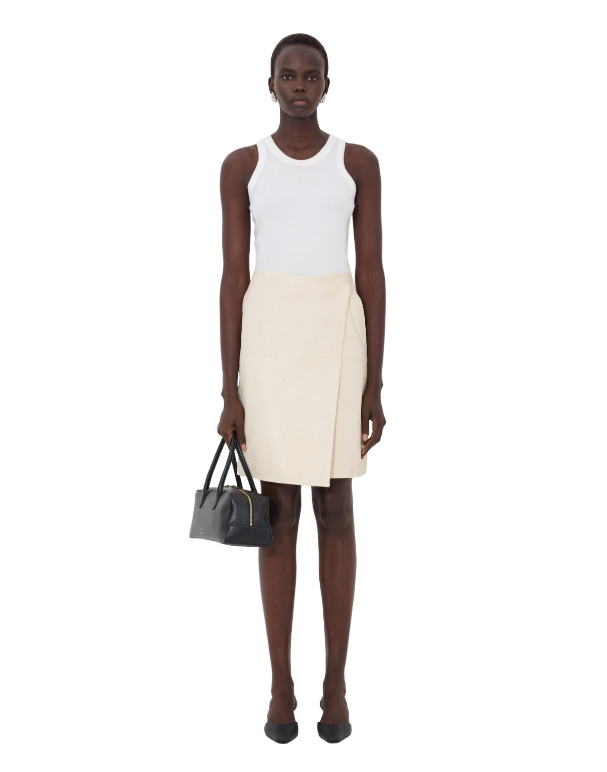 Woman wearing a white sleeveless top and beige skirt holding a black handbag on a white background