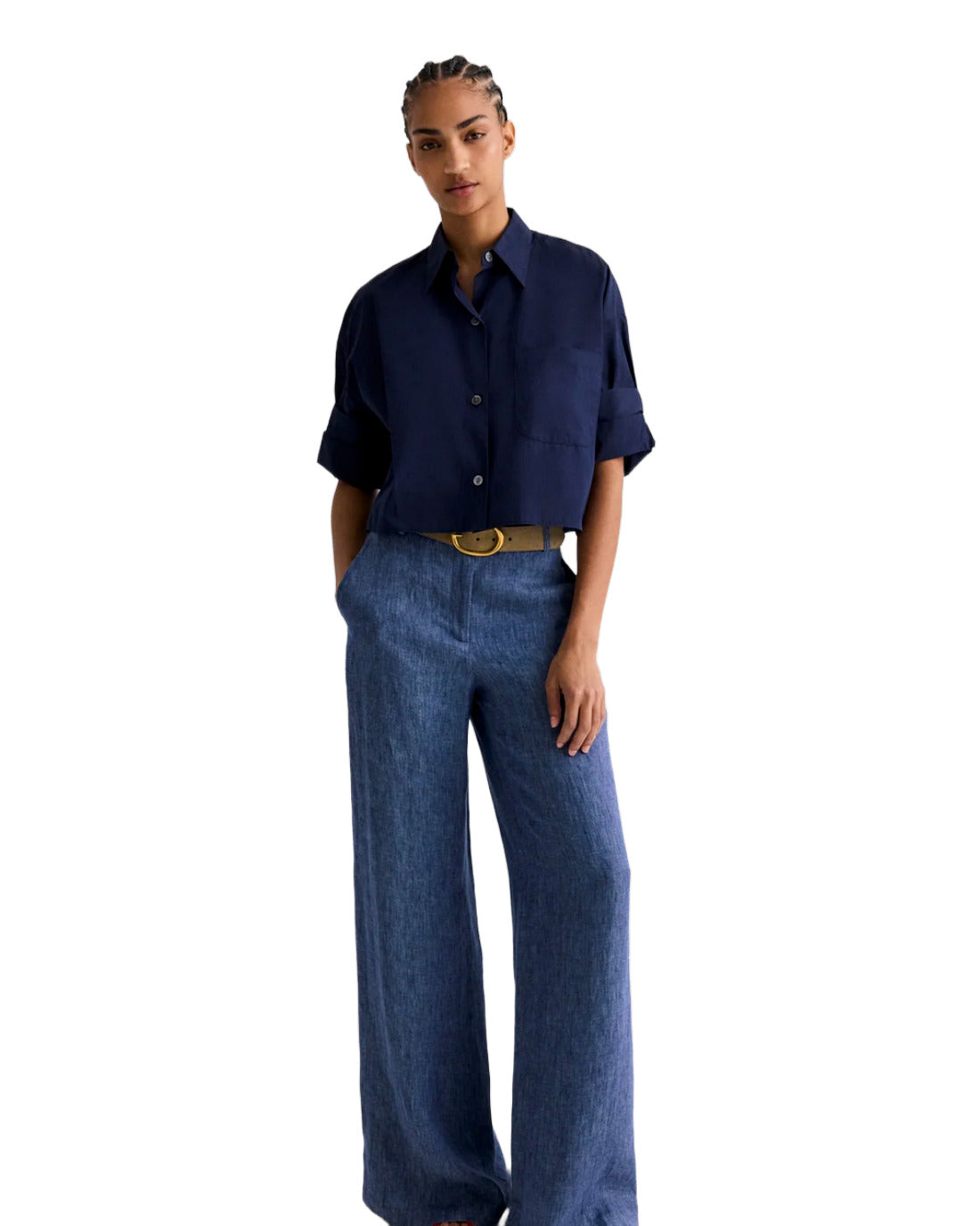 Full length view of a model wearing a navy short sleeve button front shirt with a relaxed fit and rolled cuffs, paired with wide leg blue trousers and a tan belt, standing against a white background.