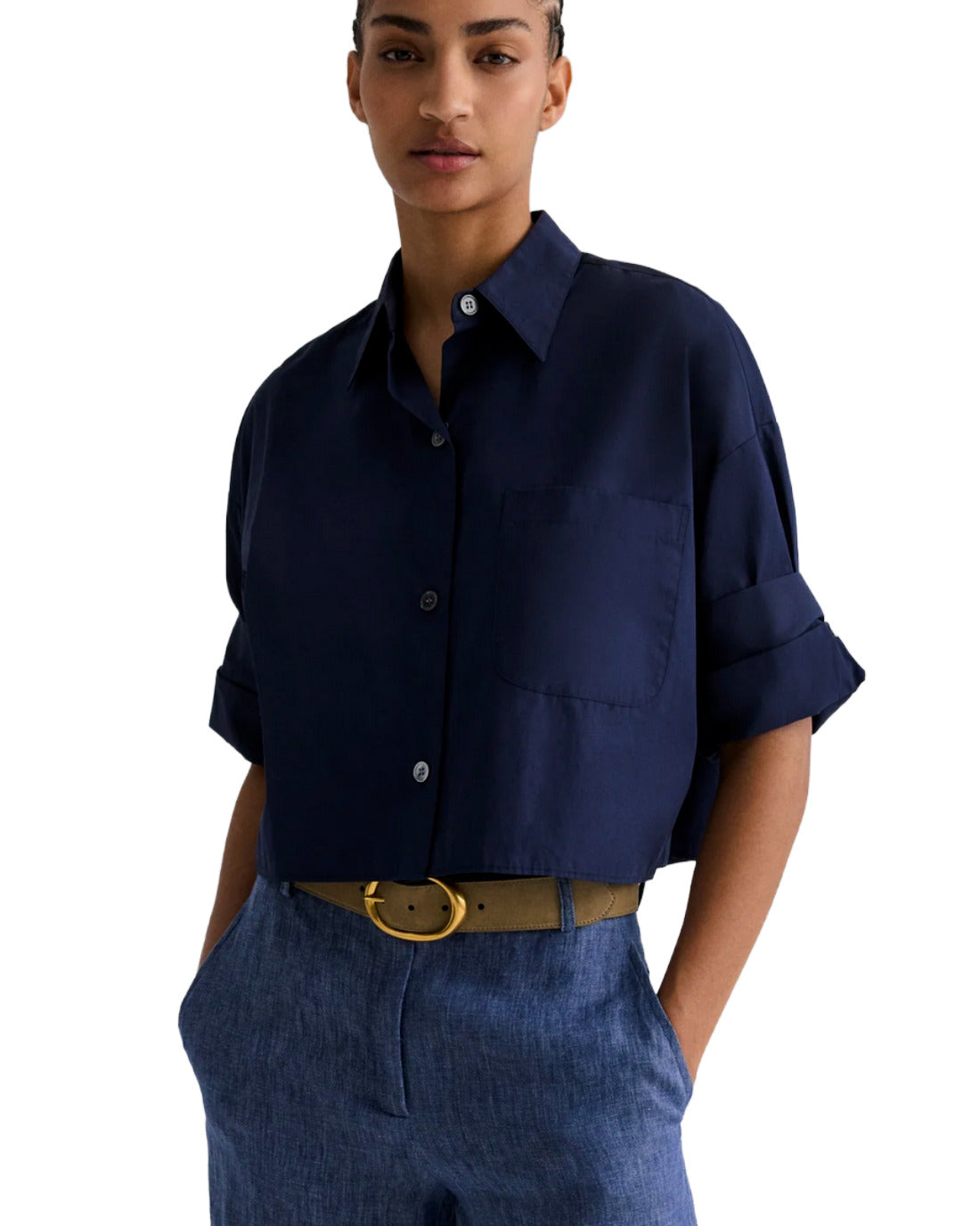 Model wearing a navy short sleeve button front shirt with a relaxed fit, chest pocket, and rolled cuffs, styled with blue trousers and a tan belt against a white background.