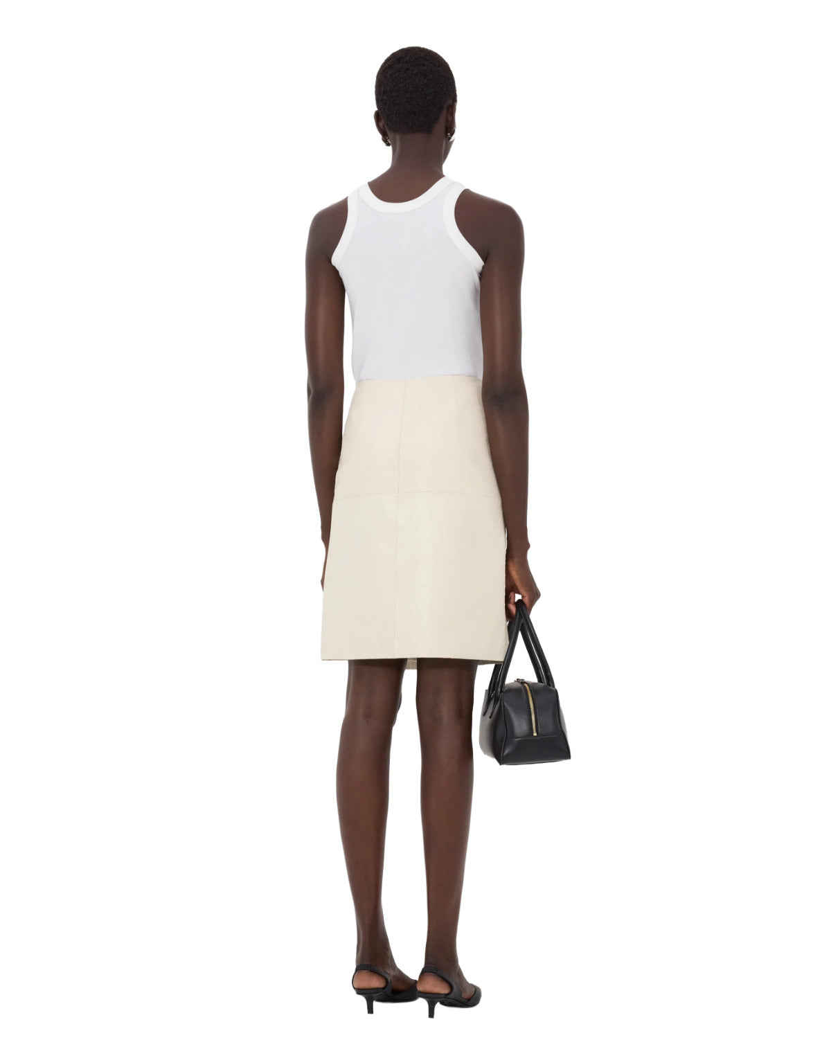 Person wearing a white sleeveless top and cream skirt holding a black handbag on a white background