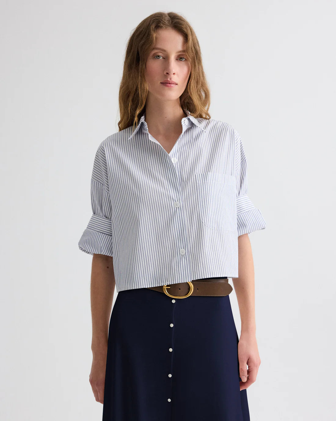 Model wearing a blue and white striped short sleeve button front shirt with a relaxed cropped fit and chest pocket, styled with a navy skirt and brown belt against a light neutral background.